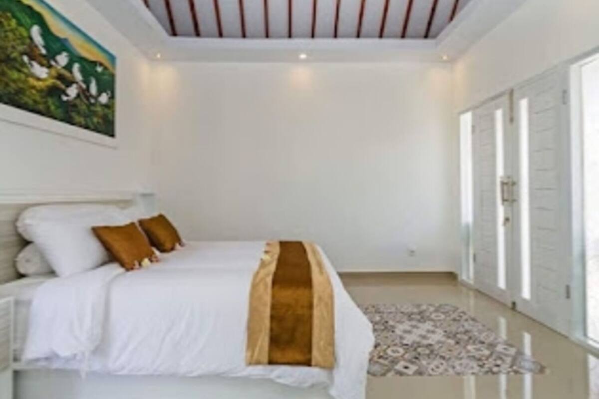 Quarto com cama, almofadas, tapete, pintura e teto com vigas de madeira.