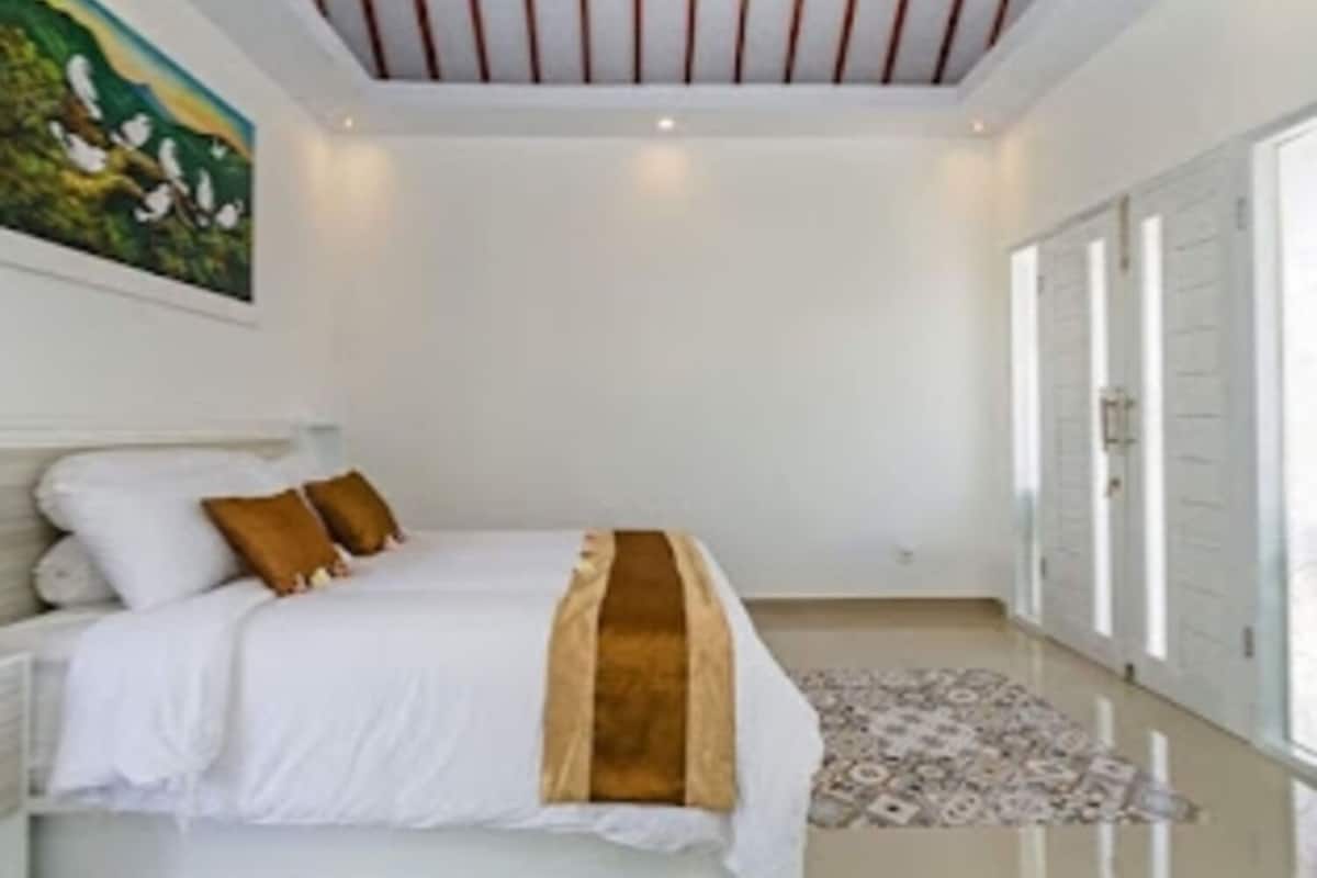 A bedroom with a bed, pillows, a rug, a painting, and a ceiling with wooden beams.