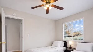 Une chambre avec deux lits, un ventilateur de plafond, une fenêtre avec des stores et un plancher en bois.