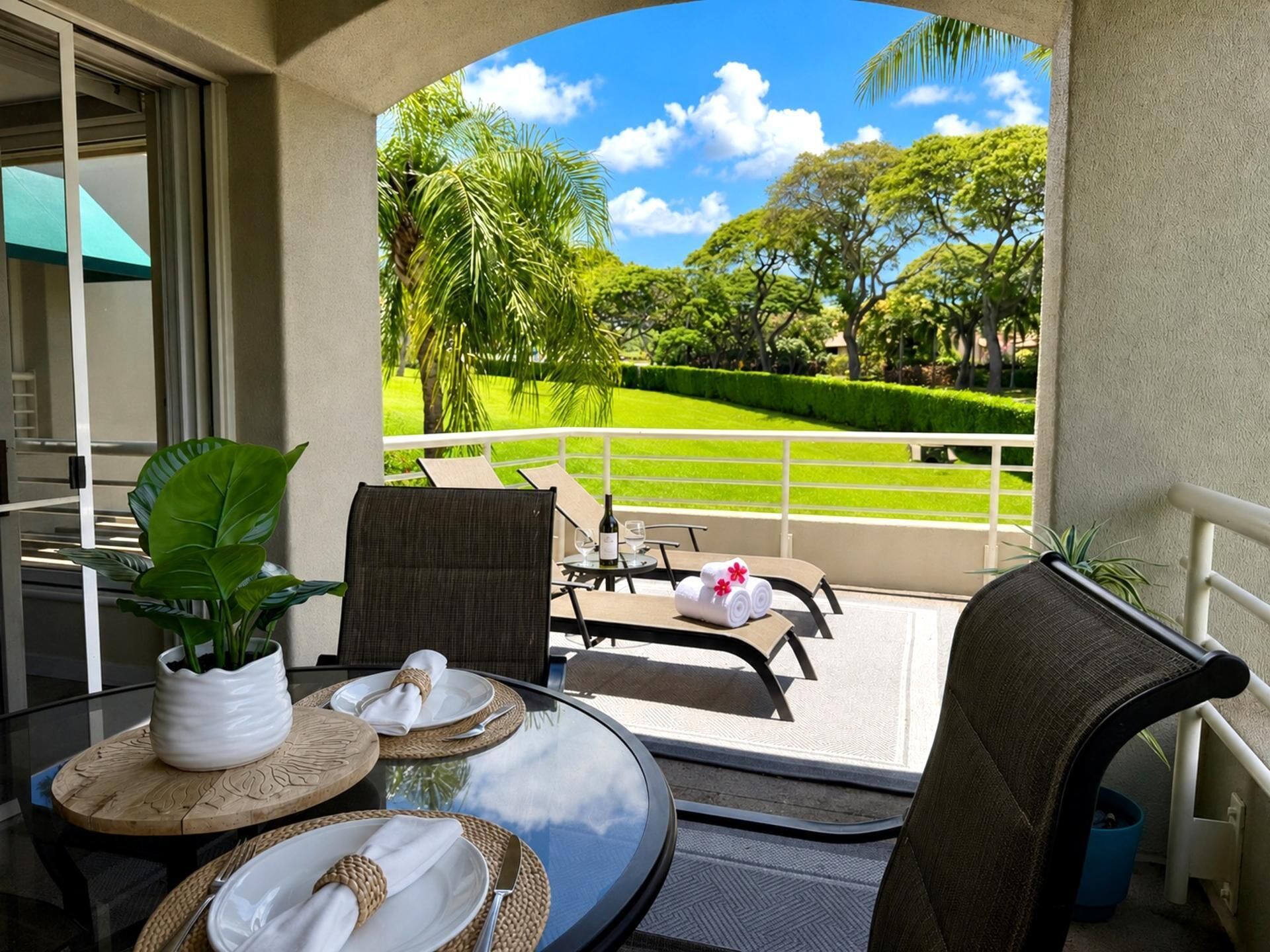 A balcony with a table set for two, a potted plant, and a view of a well-manicured lawn and trees.