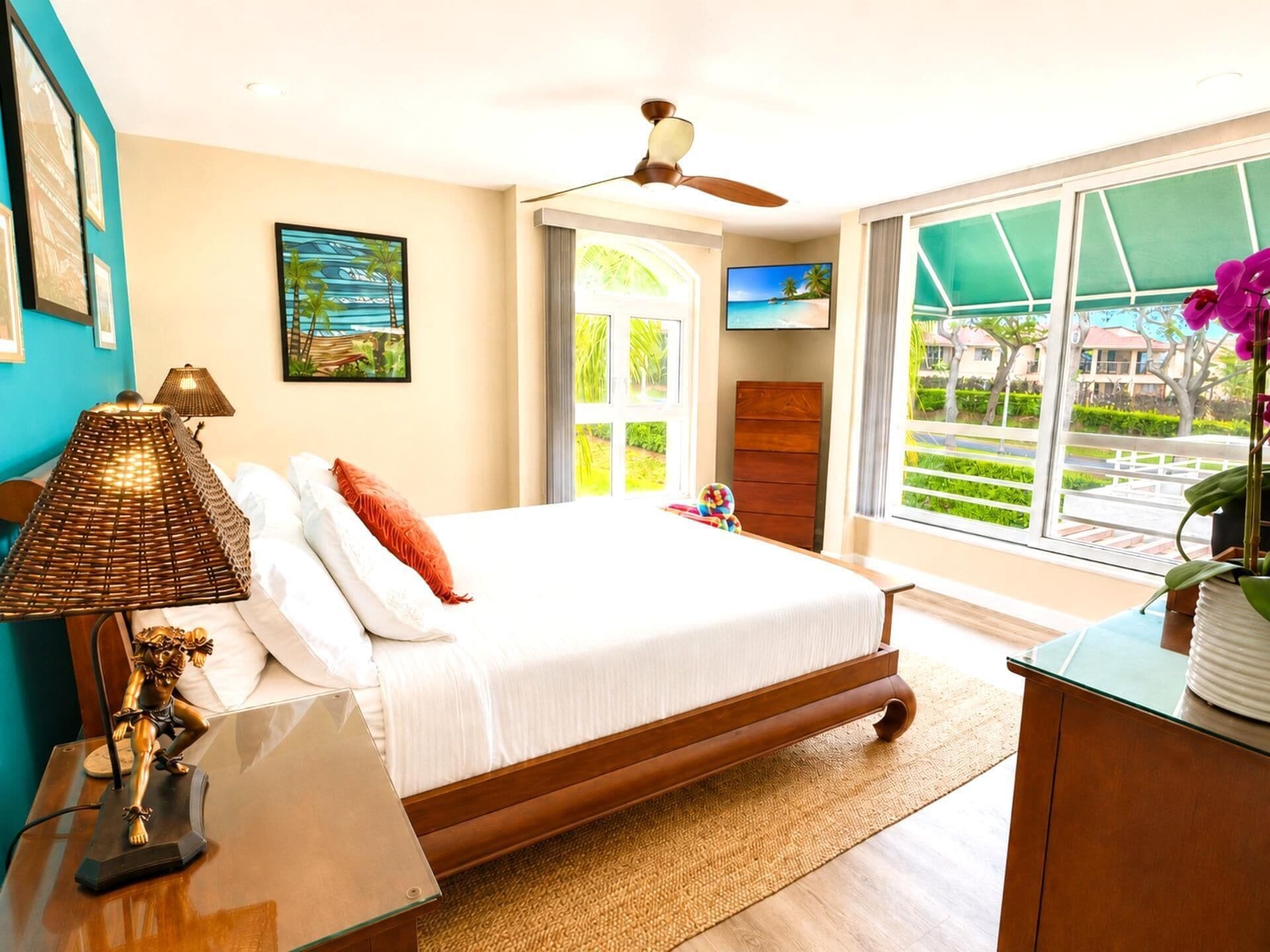 A bedroom with a bed, a desk, a lamp, a television, and a window with a view of greenery.