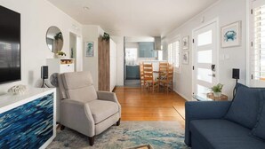A modern living room with a sofa, armchair, and a TV on a white console. A dining area with a table and chairs is visible in the background. - Golden Hour Overlook | Spa | In-Law Suite | Grill (San Diego)