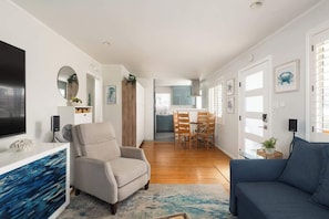 Ein modernes Wohnzimmer mit Sofa, Sessel und einem Fernseher auf einer weißen Konsole. Im Hintergrund ist eine Essecke mit Tisch und Stühlen zu sehen.