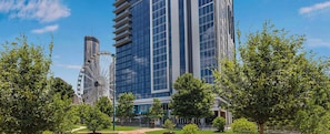 A modern building with a glass facade, a Ferris wheel in the background, and surrounding greenery.