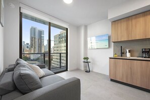 Ein modernes Apartment mit grauem Sofa, einem kleinen runden Tisch mit Pflanze, einer Küche mit Holzschränken und einem großen Fenster mit Blick auf die Stadt.