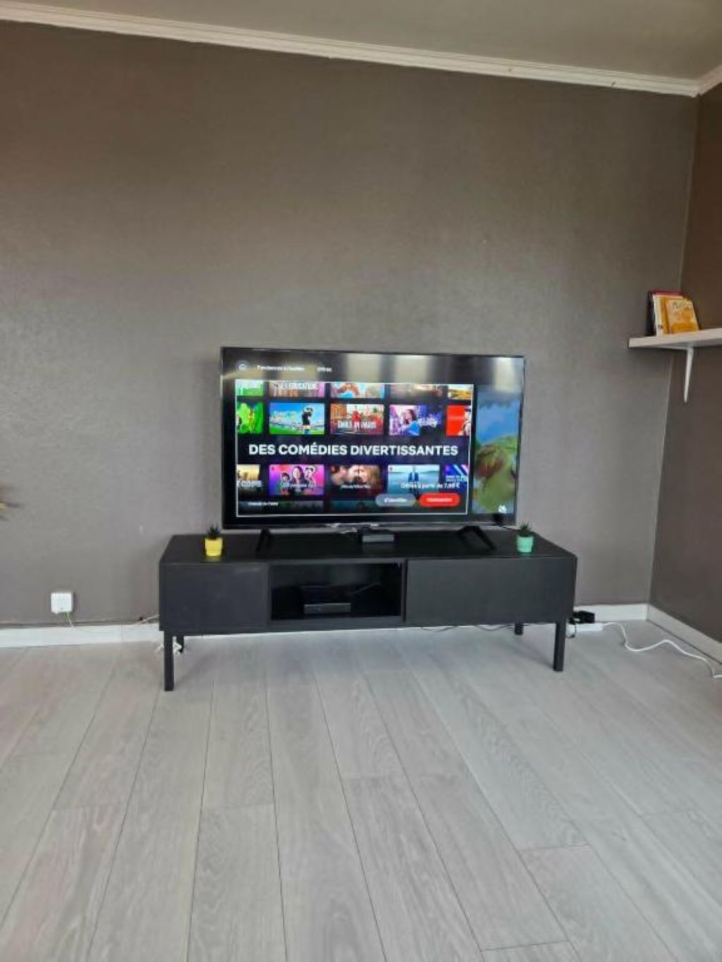 A flat-screen TV on a black stand in a room with grey walls and light wooden flooring.
