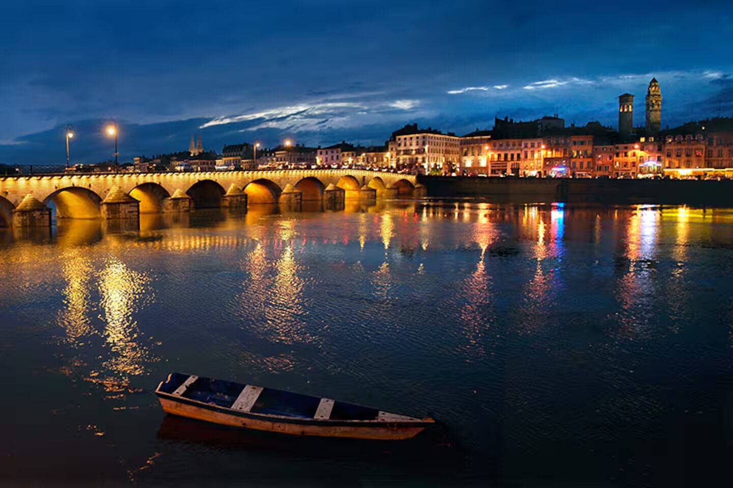 Vista nocturna de un río con un puente, una embarcación y edificios iluminados al fondo.