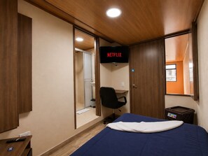 Ein Zimmer mit einem Bett, einem Schreibtisch, einem Sessel und einem Fernseher, der 'NETFLIX' anzeigt, sowie einem Fenster.