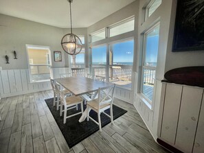 Ein Essbereich mit Holztisch und -stühlen, großen Fenstern mit Blick auf den Strand und einer hängenden Leuchte.