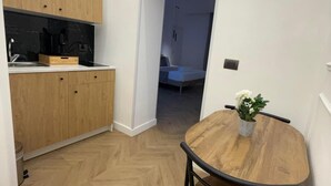 Une cuisine compacte avec des meubles en bois et un parquet en chevrons, qui donne sur une chambre à coucher équipée d’un lit et d’une table de chevet.