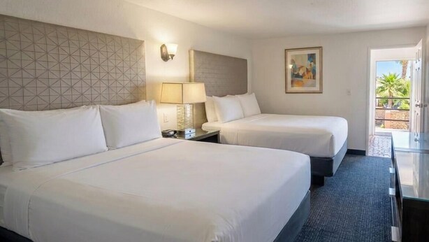 A hotel room with two beds, a nightstand, a lamp, a painting, and a view of the outdoors. - Hotel Palmas Quintero (Playas de Rosarito)