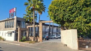A hotel with a prominent 'HOTEL' sign, a parking lot with several cars, and a large tree in front. - Hotel Palmas Quintero (Playas de Rosarito)