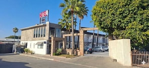 A hotel with a prominent 'HOTEL' sign, a parking lot with several cars, and a large tree in front. - Hotel Palmas Quintero (Playas de Rosarito)