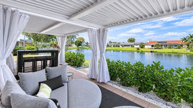 A covered outdoor seating area with a view of a lake and a house in the background. - Stylish Lake View House: Minutes to Beaches & IMG, Pet-friendly (Bradenton)
