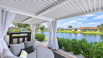 Una zona salotto all'aperto coperta, con vista su un lago e una casa sullo sfondo.