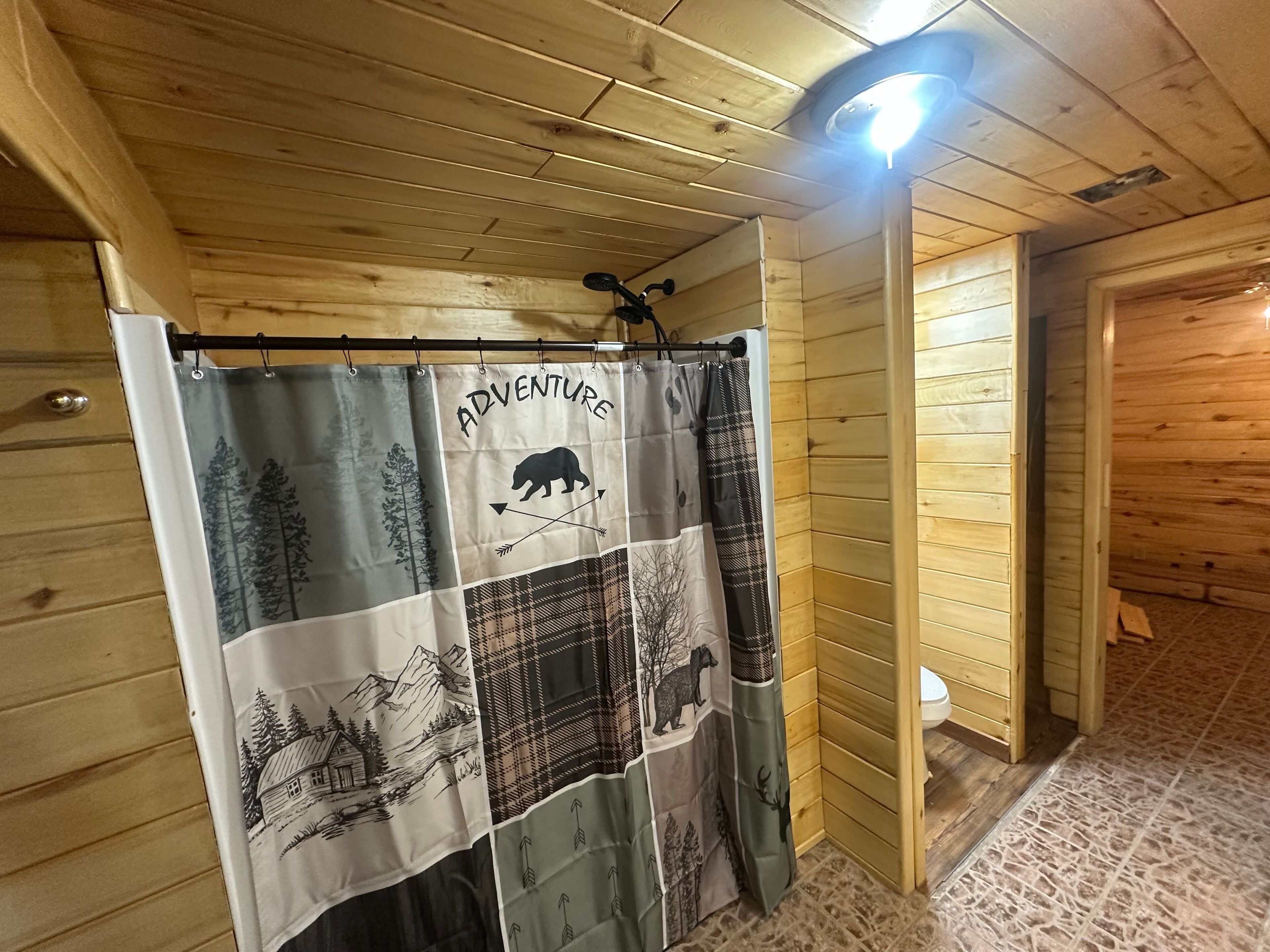 L’intérieur d’une cabane en bois, avec un rideau de douche orné d’un ours et de flèches, un luminaire au plafond et un plancher en bois.