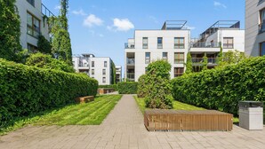 Um edifício residencial moderno com um caminho pavimentado, vegetação e um banco de madeira.