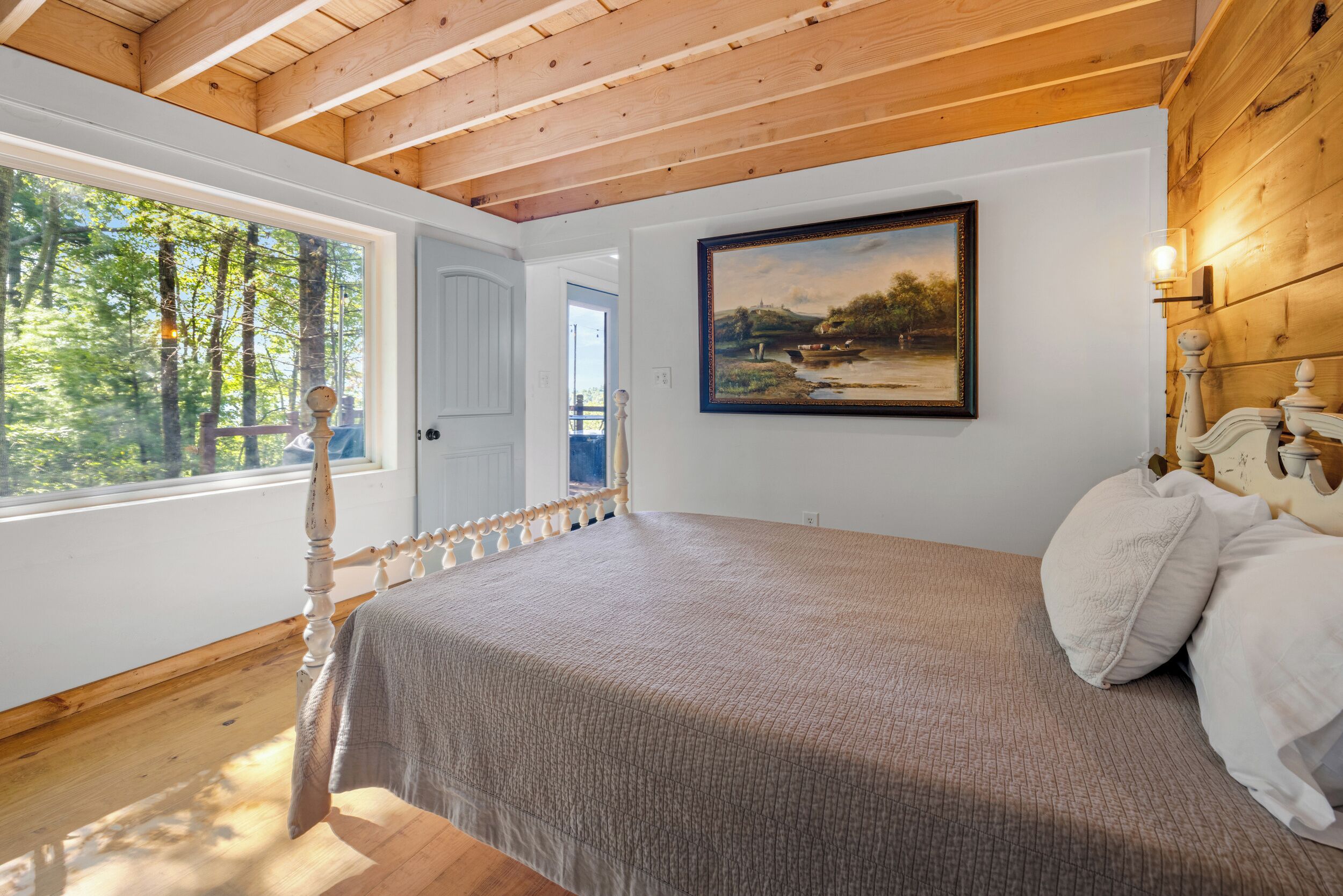 A bedroom with a large window, a bed with a headboard, and a painting on the wall.