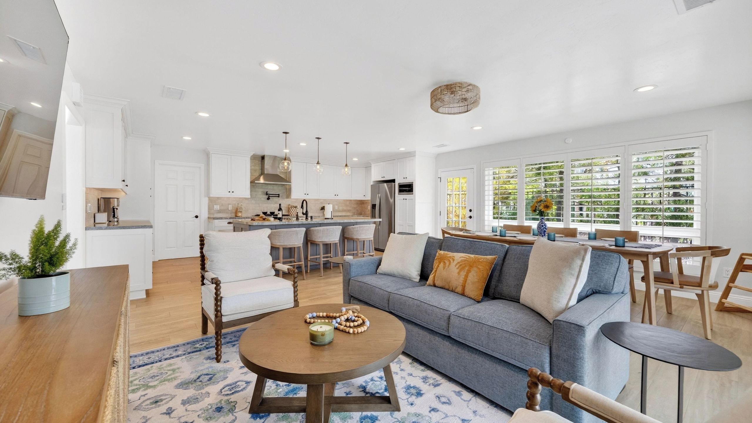A modern living room with a sofa, coffee table, and chairs, a kitchen with white cabinets, and large windows with blinds.