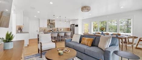 A modern living room with a sofa, coffee table, and chairs, a kitchen with white cabinets, and large windows with blinds.