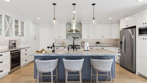 Een moderne keuken met witte kastjes, een centraal eiland met barstoelen, een roestvrijstalen koelkast en hangende lampen.