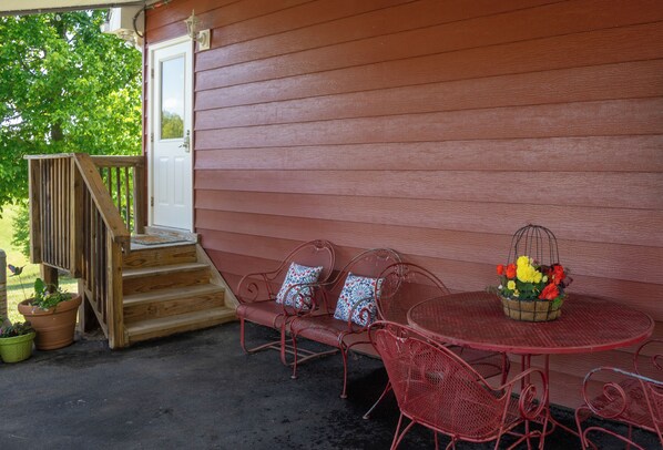 A wooden porch with a red metal table and chairs, a small step leading to a white door, and potted plants.