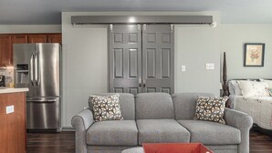 Ein Wohnzimmer mit grauem Sofa, einem roten Tablett-Tisch und einer Küchenzeile mit einem Edelstahl-Kühlschrank.