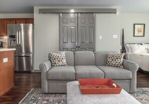 Una sala de estar con un sofá gris, una mesita roja y una cocina con un refrigerador de acero inoxidable.