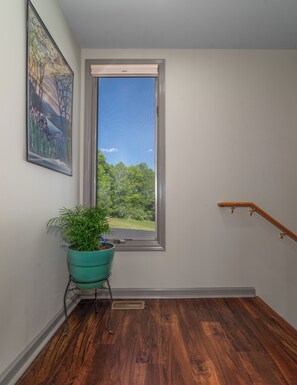 Une pièce dotée d’un plancher en bois, d’une fenêtre donnant sur un espace verdoyant, d’une plante en pot et d’un tableau accroché au mur.