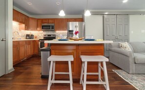 Cocina con armarios de madera, un refrigerador de acero inoxidable, un microondas y una cafetera. Hay dos taburetes blancos y un sofá gris en la sala.
