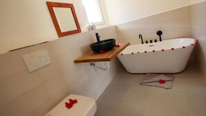 A bathroom with a white bathtub, a toilet, and a wooden shelf with a black basin. - Family Room (Fumba)