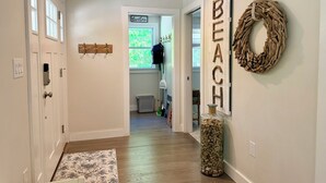 Un pasillo con un perchero de madera, un florero de vidrio con trozos de madera y una corona decorativa en la pared.
