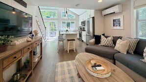 Una cocina moderna con una isla central, una zona de comedor con bancos, una sala de estar con sofá y ventanas amplias.