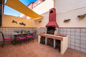 Eine Terrasse im Freien mit Ziegelboden, einem Tisch mit Stühlen, einem Grill und einem gelben Sonnensegel.