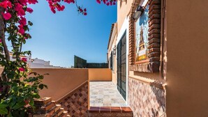 Un estrecho pasillo exterior con suelo de baldosas, pared de ladrillo y una puerta de metal.