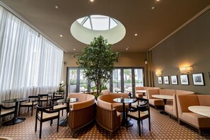 Ein stilvoller Lounge-Bereich mit runden Tischen, bequemen Sitzgelegenheiten, einem großen Baum und einem Oberlicht.