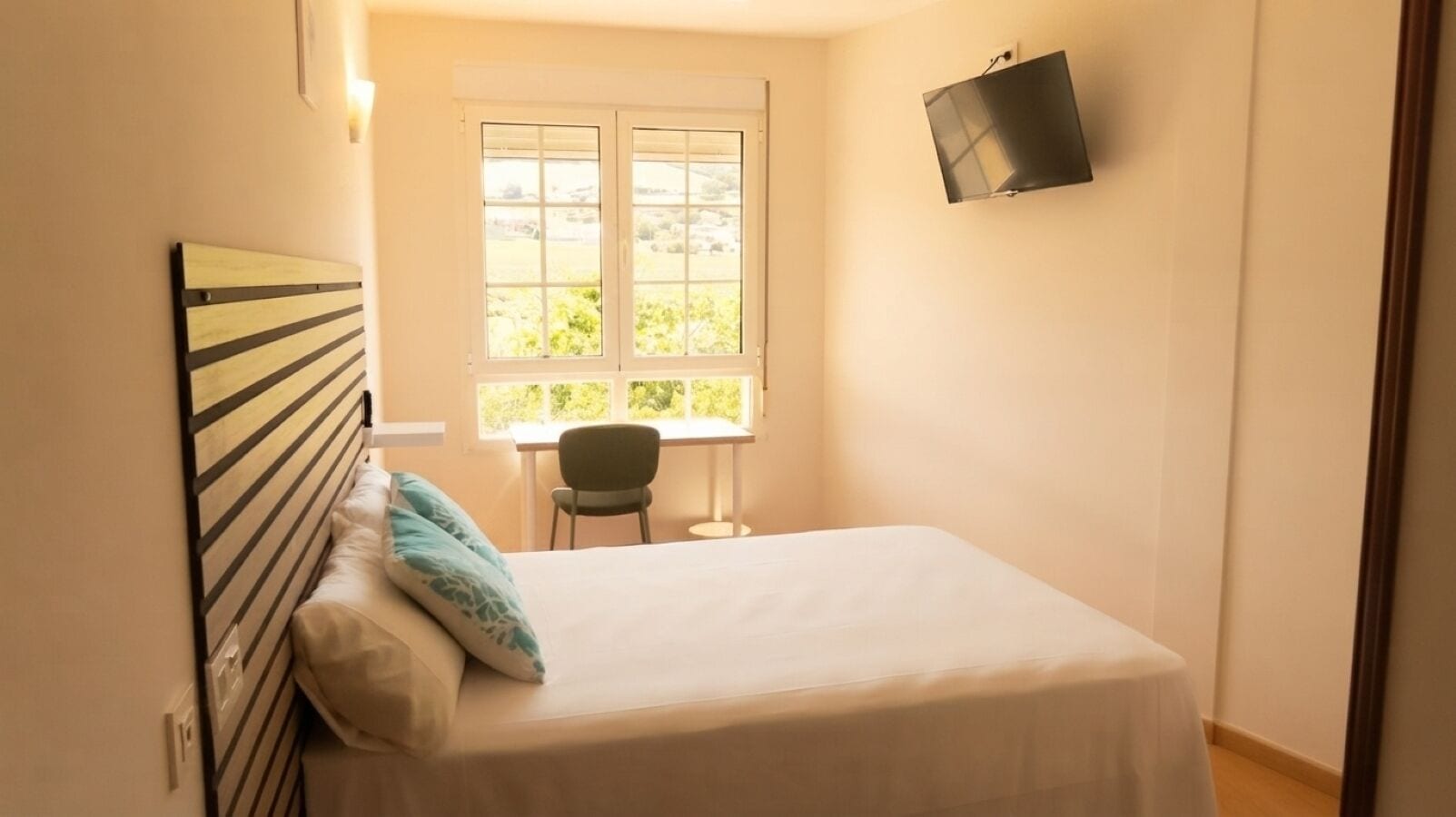 A bedroom with a bed, a chair, a window, and a TV mounted on the wall.