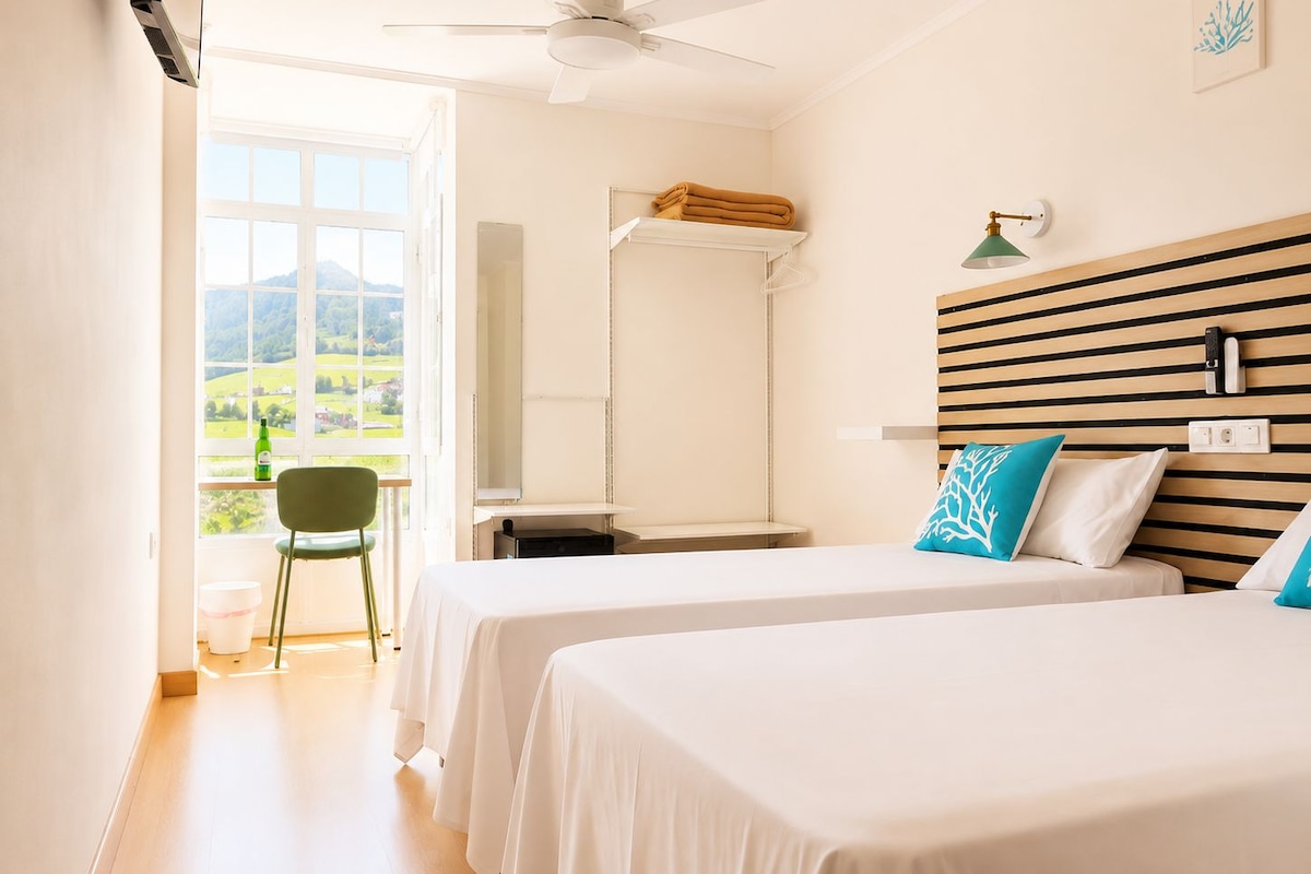 Habitación de hotel con dos camas, un escritorio, una silla, un ventilador de techo, una ventana con vistas al campo y un estante en la pared.