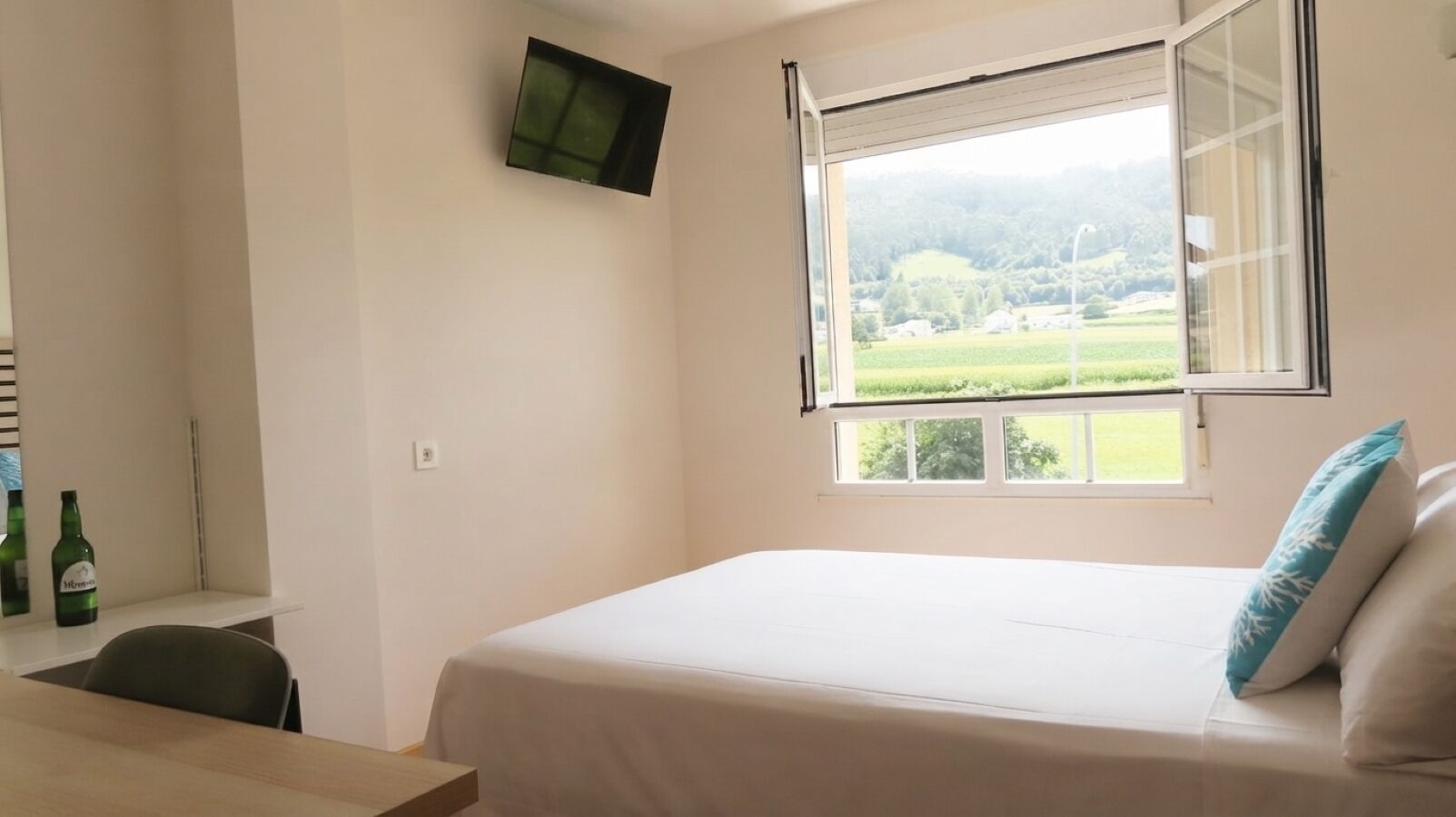 Un dormitori amb un llit, una taula de treball, una cadira, una finestra amb vista a vegetació i un televisor de pantalla plana fixat a la paret.