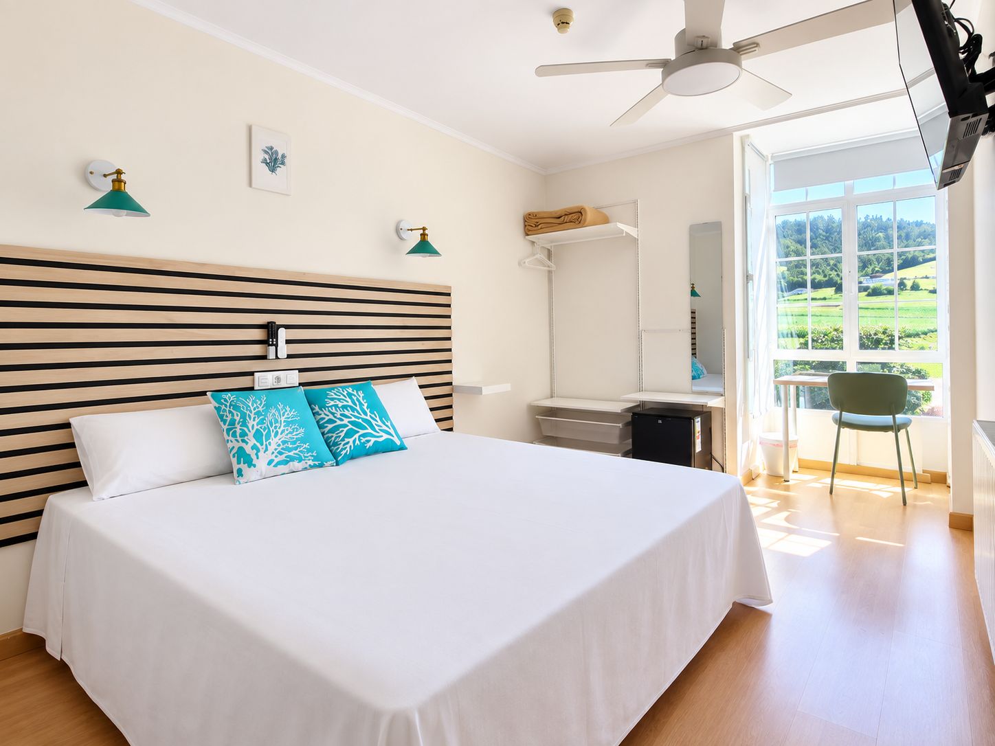 A hotel room with a bed, a desk, a chair, and a view of greenery outside.