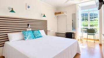 A hotel room with a bed, a desk, a chair, and a view of greenery outside.