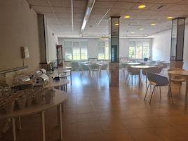 Un comedor amplio con varias mesas y sillas, una barra de buffet con platos y vasos, y ventanas grandes que dejan entrar luz natural.
