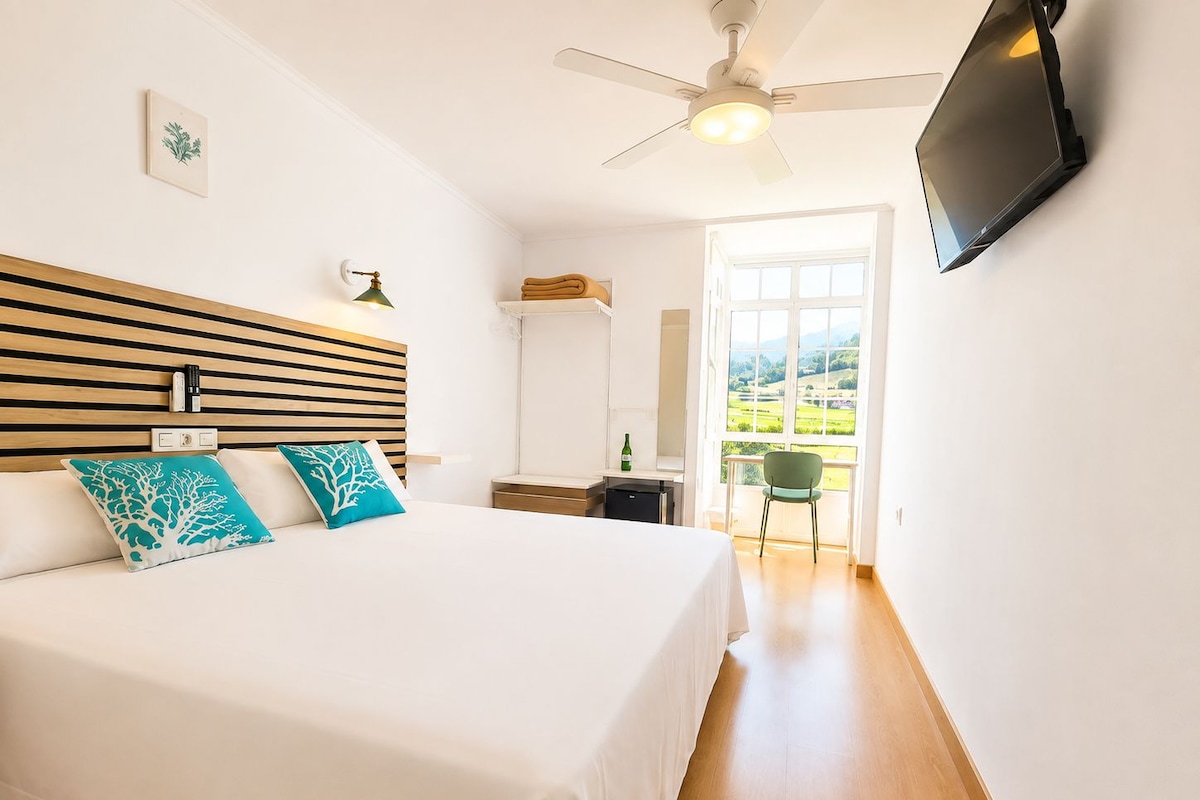 Un dormitorio con cama, escritorio, silla, televisor y vista al exterior.