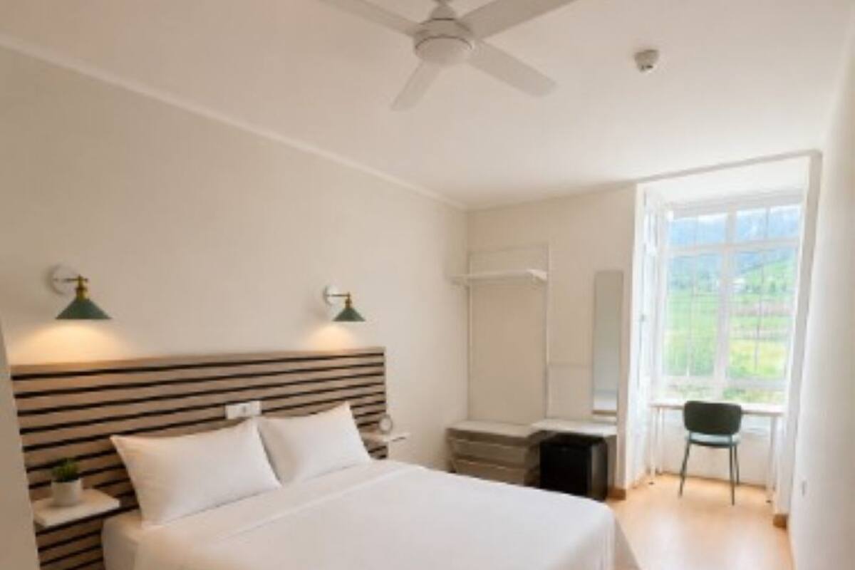 A bedroom with a bed, a desk, a chair, a ceiling fan, and a window.