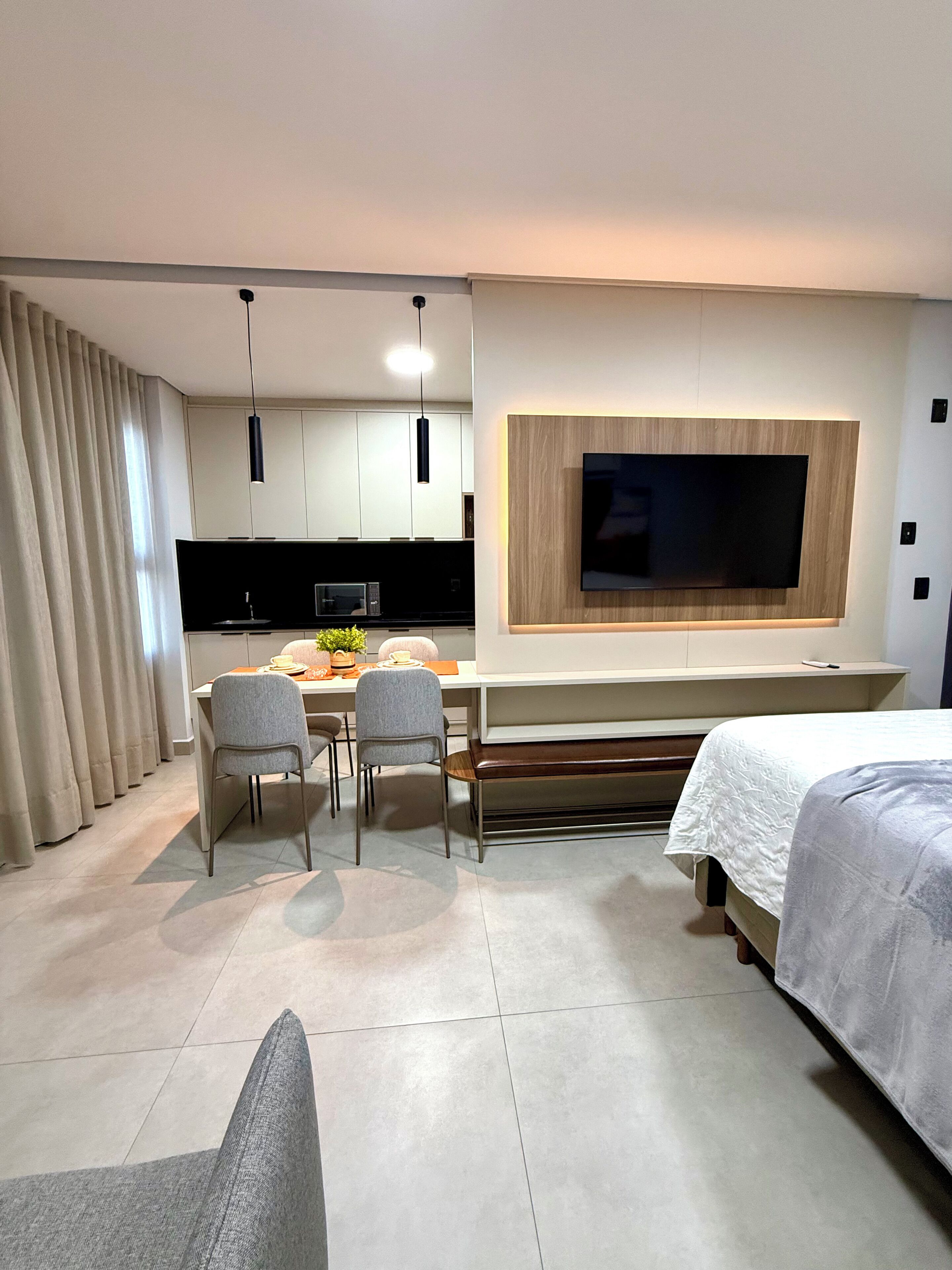Una habitación de hotel moderna con zona de comedor, un televisor instalado en un panel de madera y una cama con edredón blanco.