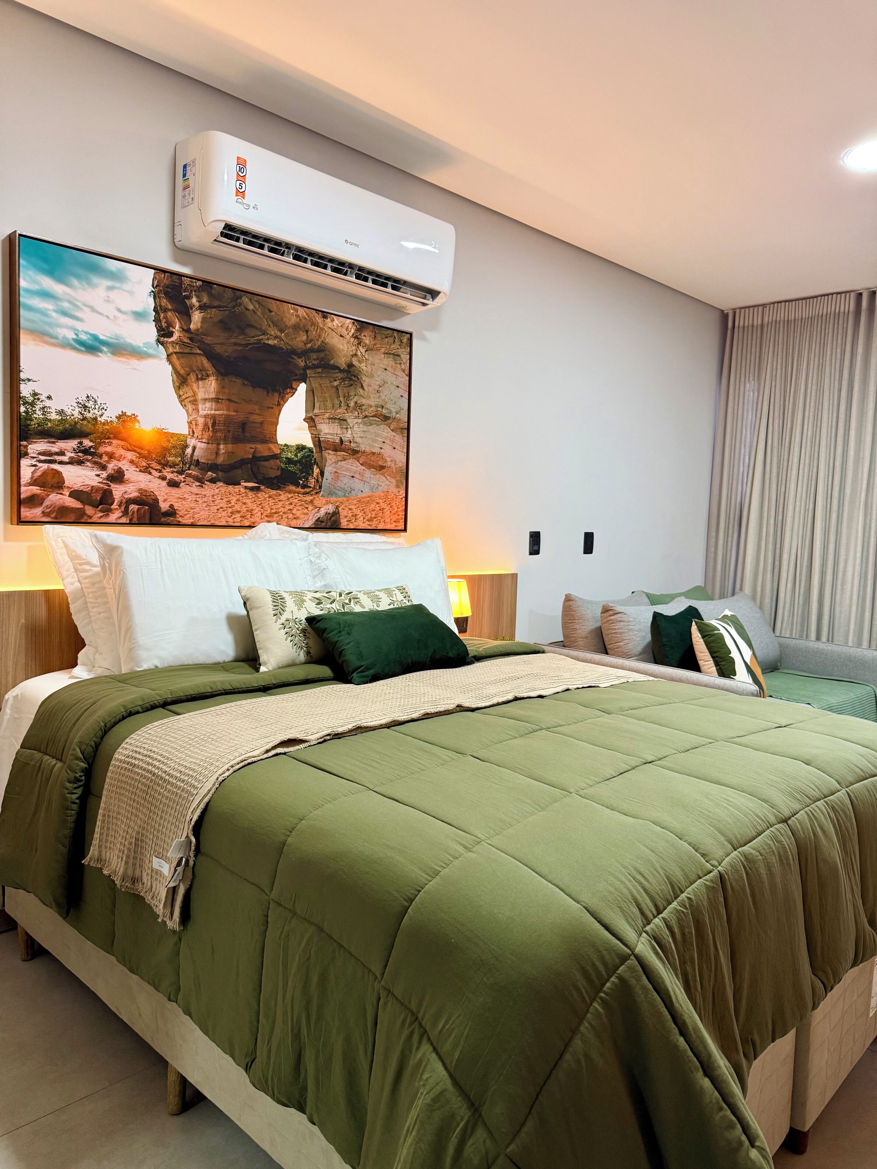 Uma cama bem arrumada com um edredom verde, um ar condicionado fixado na parede, um quadro com uma formação rochosa e uma janela com cortinas.