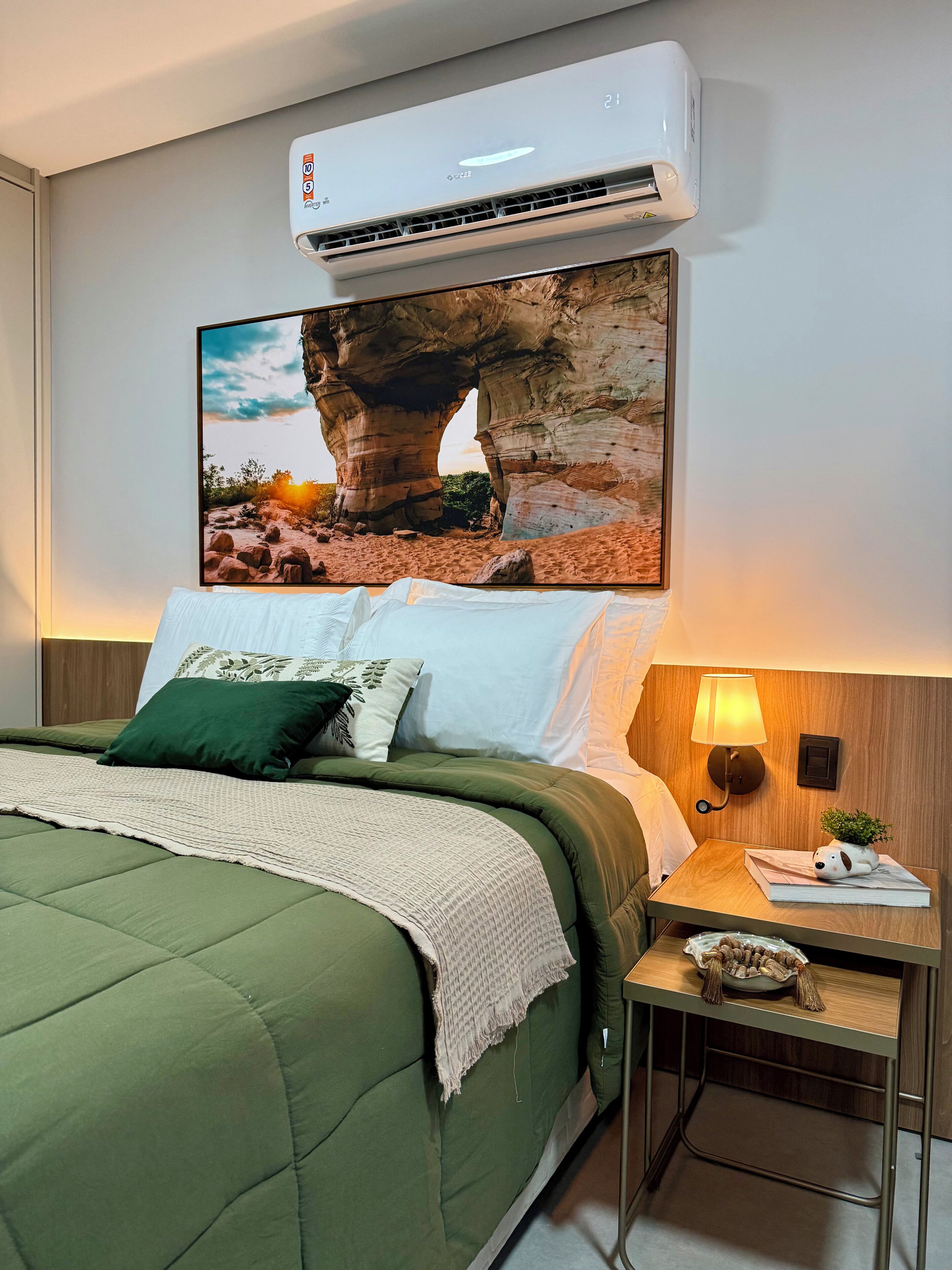 A hotel room with a bed, a nightstand, a lamp, a wall-mounted air conditioner, and a large framed picture of a rock formation.