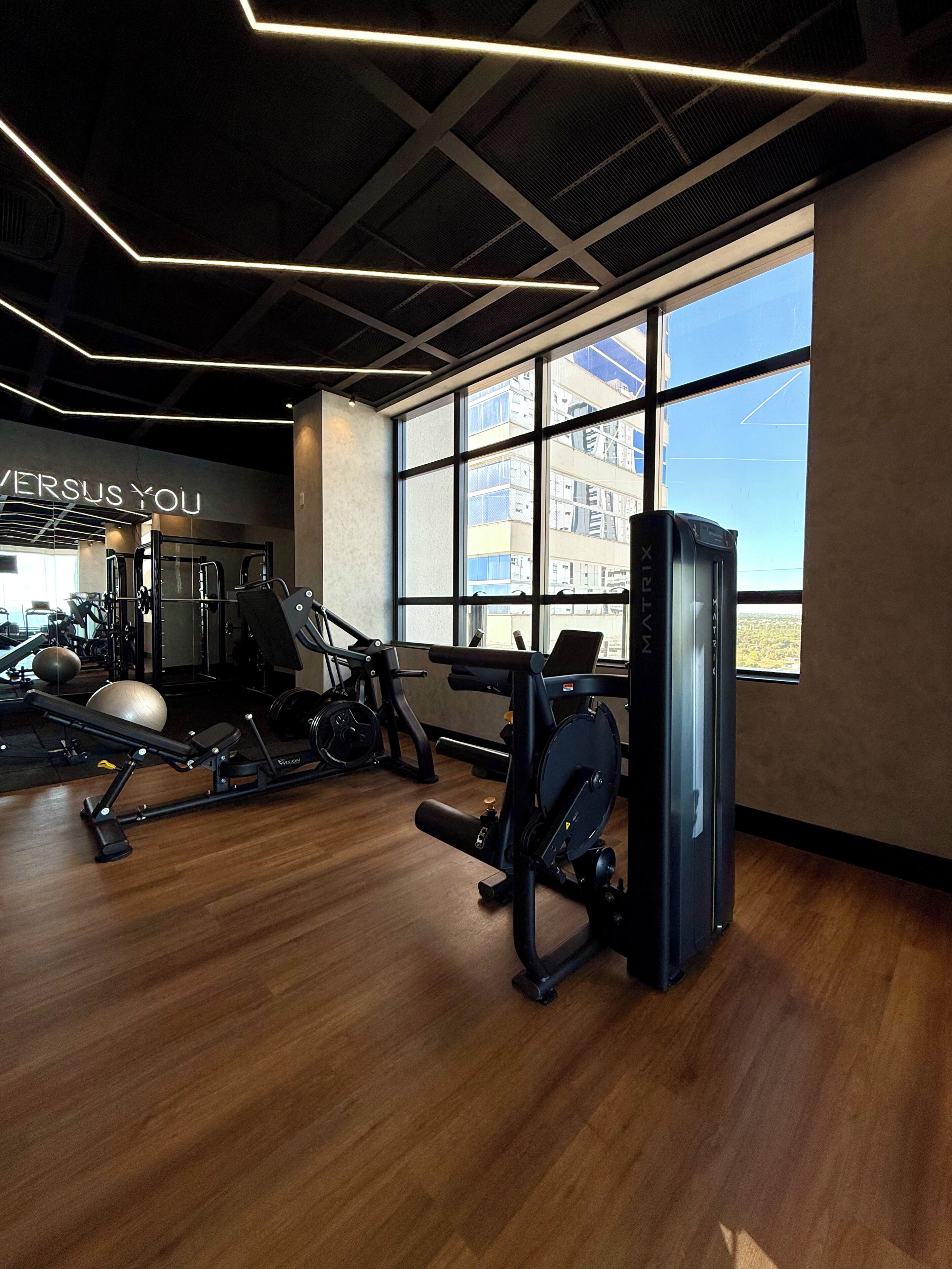Uma academia com equipamentos de ginástica, uma janela grande e um letreiro na parede com a inscrição 'VERSUS YOU'.