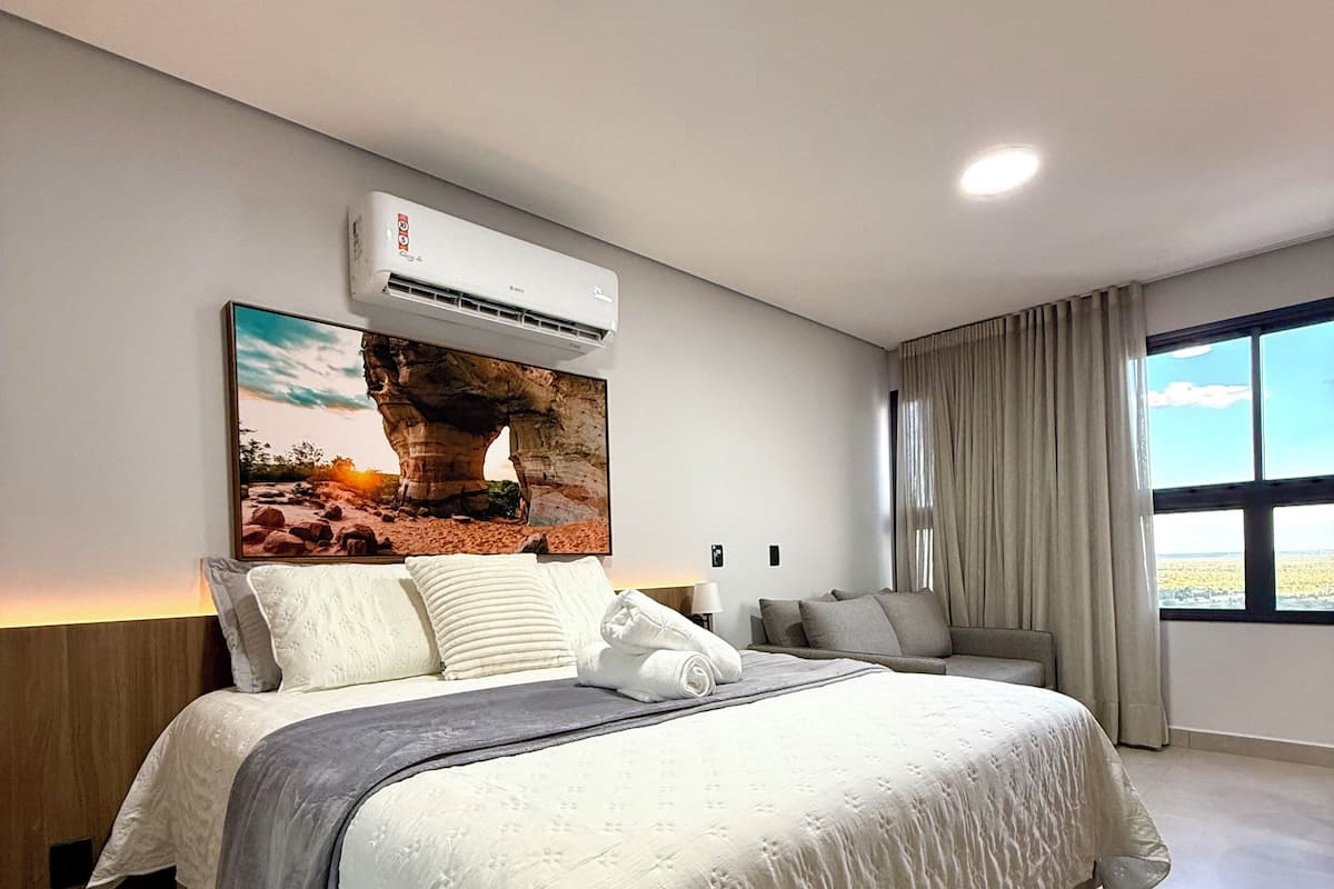 Ett modernt hotellrum med en stor säng, en väggmonterad luftkonditioneringsanläggning, ett stort fönster med gardiner och ett konstverk som hänger på väggen.