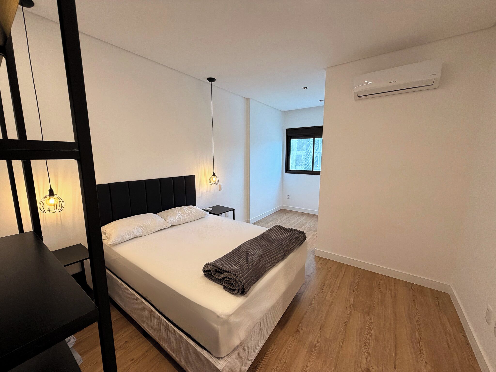 Une chambre à coucher comprenant un lit, des tables de chevet, un luminaire suspendu, une fenêtre et un appareil de climatisation.